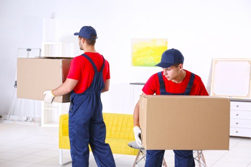 Two removal workers in matching navy blue caps, red shirts, and blue overalls are engaged in a house moving procedure inside a bright, modern living room. The worker on the left, standing with his back to the camera, is carrying a medium-sized cardboard box while wearing white gloves. The other worker, on the right, is kneeling or bending slightly, handling a larger cardboard box, also wearing white gloves. In the background, there is a yellow sofa positioned against a white wall, which has abstract artwork hung above it featuring yellow and green tones. To the right, a white chest of drawers with minimalist design is visible, and to the left, a tall white shelving unit holds several decorative objects and storage boxes. The room is well-lit with natural light, creating a spacious and clean environment suitable for residential removals in the London area, including the Watson postcode region. The scene captures a professional moving team efficiently organizing household items with cardboard boxes, ready for transportation, reflecting reliable removals services typical for houses in London.