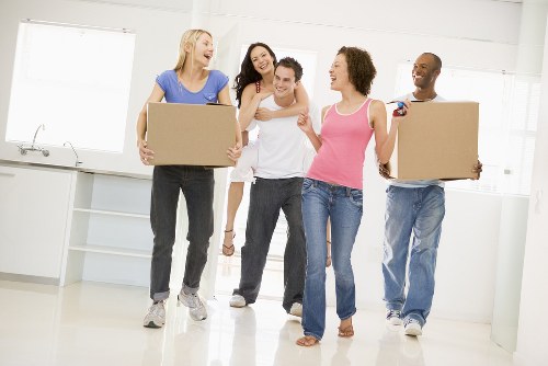 A group of five diverse individuals in a bright, modern residential kitchen, preparing for a house move. Two women and one man are carrying large cardboard boxes, with one woman in a blue top and grey jeans holding a box on her right side, and another woman in a pink top and blue jeans holding a box on her left side. Behind them, a woman with dark hair in a white top and grey trousers is smiling and making a gesture of encouragement towards the others. A man with a shaved head and dark skin, dressed in a striped shirt and jeans, stands to the side holding a box. The kitchen features white walls, large windows with abundant natural light, and minimalist cabinetry, suggesting a clean, contemporary home environment typical of London or nearby areas. The scene captures the collaborative effort of a professional removals team, likely from Watson Removals London, in the process of loading or unloading household belongings during a move within the local area, highlighting the practical aspects of house removals and moving services in the UK.