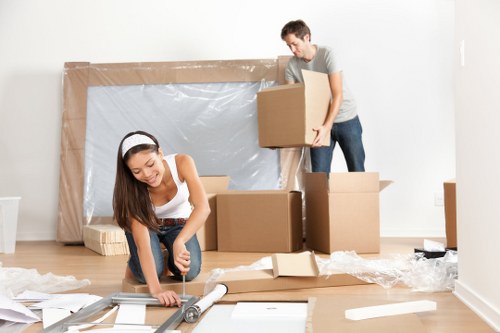 A young woman with long brown hair, wearing a white sleeveless top and blue jeans, is kneeling on a light wooden floor as she uses a utility knife to cut packaging materials on a paper-wrapped parcel. Behind her, a young man with short dark hair, dressed in a grey t-shirt and dark jeans, is lifting a medium-sized cardboard box, preparing to load or move it. The room appears to be an interior space with plain white walls and is being prepared for a house move, as indicated by the presence of several stacked and scattered cardboard boxes, some open and others sealed, along with packing materials such as bubble wrap and crumpled paper. In the background, a large piece of furniture or packaging wrapped in plastic and cardboard is leaning against the wall, possibly for protection during the move. The scene suggests a professional removals process taking place, with attention to careful packing and handling, typical of services offered by Watson Removals London in Upton Park, close to the E13 postcode area, in an environment of a residential property ready for relocation within or from the area.
