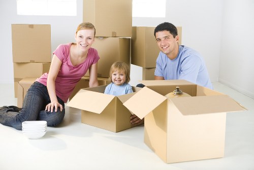 A family with two adults and a young child are inside a well-lit, empty residential room, surrounded by multiple cardboard boxes in various stages of packing or unpacking. The woman is seated on the floor, wearing a pink short-sleeved top and jeans, smiling at the camera. The young girl, seated in a partially open box, has blonde hair and is dressed in a light blue shirt, also smiling. The man, wearing a light blue T-shirt, is kneeling next to a box, smiling at the camera. Behind them, several closed and open cardboard boxes are stacked against white walls, indicating a move or packing process typical of house removals in the London area. The scene conveys a sense of preparation for a household relocation, highlighting the use of sturdy cardboard cartons and an organized indoor environment, characteristic of professional removals services like Watson Removals London.