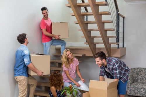 A group of five individuals, two men and three women, are engaged in unpacking and moving cardboard boxes inside a modern, well-lit house with a staircase, which may be part of a house removal or relocation process in Colney Hatch. One man dressed in a red t-shirt and jeans is standing on a lower step of the staircase, holding a medium-sized cardboard box, while another man in a blue checkered shirt is crouched nearby, assisting with a box. A woman in a pink and white striped top is standing in the middle, smiling and holding a clear plastic bag, with another woman beside her, seated and holding a small potted plant on a table or countertop. The scene includes visible household furniture such as a floral-patterned armchair, and the staircase is made of wood with metal railings, set against a white wall. The natural lighting accentuates the casual, organized activity typical of house removals in the Colney Hatch area, with the group proceeding in a friendly, collaborative manner to unload or pack belongings in the interior space.