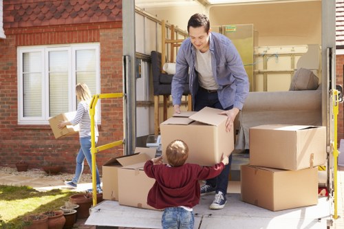 A professional man wearing casual clothing is loading cardboard boxes into a white removals van parked in front of a residential house with a red brick exterior. The van's rear doors are open, revealing the interior with more packed boxes and a grey upholstered sofa. A young girl, dressed in a striped long-sleeve top and jeans, is walking away from the van carrying a box towards the house, which has white-framed windows and a small garden area with potted plants. The scene suggests a house removal or relocation process taking place on a clear day, with natural daylight illuminating the scene. The presence of multiple packed boxes indicates an organized move, and the partially visible outdoor environment and residential setting subtly connect to local London moving services, such as those provided by Watson Removals London.