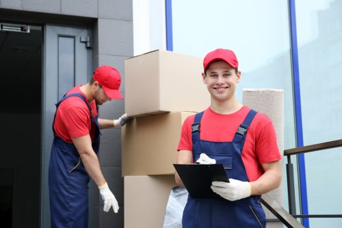 Two removal workers dressed in red caps and blue overalls are engaged in a house moving task outside a modern residential building, which features large glass windows with blue framing and a dark grey entrance door. One worker, positioned on the right, is smiling at the camera while holding a clipboard and wearing light-colored gloves, appearing confident in his role. The other worker, on the left, is focused on stacking three large cardboard boxes, each with smooth brown surfaces, against the building's exterior wall. The boxes vary in size and are arranged vertically, with the smallest at the top. Behind the workers, to the right, a textured beige carpet or rug leans against the window. The scene is well-lit, likely during daytime, with natural outdoor lighting highlighting the clean, professional environment associated with Watson Removals Londonâ€™s services in Brompton, as part of their comprehensive residential removals process.
