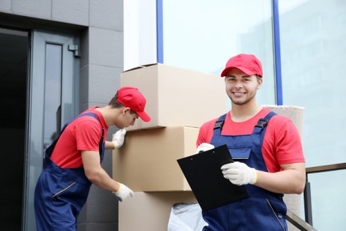 Two professional movers from Watson Removals London are working outdoors near a modern residential building in London, with one mover smiling at the camera while holding a clipboard, and the other focused on securing a large cardboard box. The mover with the clipboard is wearing a red cap, a red T-shirt, and navy blue overalls, while the other is dressed similarly, both wearing white gloves. They are positioned beside a stack of three cardboard boxes, which vary in size and are taped securely, ready to be moved. In the background, a glass window reflects part of the street scene, and a dark-colored door provides access to the building. The environment appears to be on a residential street in London, possibly near the postcode inferred from the page URL, with natural lighting suggesting daytime. The scene depicts typical house removal activity, with the focus on packaging and handling household items, demonstrating professional relocation services tailored to homes in the London area.