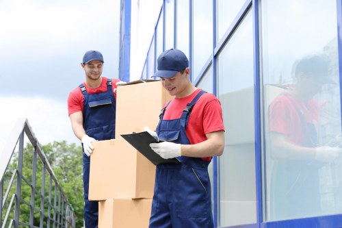 Two removal workers in red shirts, blue overalls, and matching caps are pictured outdoors during a house move. One worker, standing closer to a modern glass building with reflective windows, is holding a clipboard and appears to be inspecting or organizing paperwork, wearing white gloves. The other worker stands behind him, carrying two stacked cardboard boxes, which are light brown with visible flaps and sealed edges. The background includes a metal railing on the left side and a cloudy sky overhead, indicating an overcast day. The scene is set on a residential or commercial premises, suggesting an organized approach to an ongoing relocation process. The setting subtly hints at a professional removals service operating around the London area, consistent with Watson Removals London's offerings for house and office moves, and highlights the careful handling of household items and the use of protective clothing and equipment during the loading process.