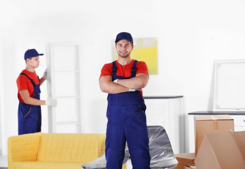 A professional removal worker stands confidently in a well-lit, modern residential interior during a house move in White City, London. He is dressed in a navy cap, red T-shirt, and blue overalls, with arms crossed and a friendly expression. In the background, another removal staff member, also in uniform, handles a white shelving unit. The room features white walls, a yellow upholstered sofa in the foreground, and a light-colored wooden sideboard. Several moving boxes, including cardboard cartons and clear plastic wrapping, are stacked around the room, indicating an active packing process. The environment appears clean and organized, with natural light illuminating the scene, emphasizing the careful handling of household items. The setting reflects typical elements of a house removal service in White City, with focus on the furniture, boxes, and the professional presence of Watson Removals London, ensuring the image is relevant and detailed for a house relocation service webpage.