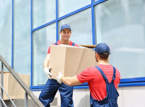 A male removal worker wearing a blue cap, red t-shirt, and blue overalls is smiling while handing a medium-sized cardboard box to a colleague outside a modern building with large blue-framed glass windows. The colleague, whose back is to the camera, is also dressed in a red t-shirt and blue overalls, and is reaching out to receive the box. The setting appears to be a residential area with a metal staircase visible on the left side, and the building's glass facade reflects the surrounding environment. The scene depicts a professional move, highlighting the careful handling of household items such as cardboard boxes, with the outdoor lighting suggesting daytime in Hounslow. This image exemplifies the services offered by Watson Removals London, supporting local house relocation efforts with efficient team collaboration.