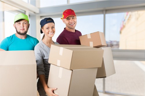 A group of three professional house movers from Watson Removals London, dressed in casual clothing and colourful caps, are standing indoors in a well-lit, modern residential space with large windows showing a view of a street in Watson or nearby London areas. They are smiling and holding several large, plain cardboard boxes, ready for loading or unloading. The boxes are stacked and held carefully, showing a range of sizes with brown kraft paper exteriors and visible packing tape. The background features a bright, clean interior with light-colored walls and a tiled floor, indicating a typical home environment in the London suburbs. This scene depicts careful handling of household items during a house relocation, highlighting the professional service provided by Watson Removals London in the area, focusing on household furniture and personal belongings being prepared for transportation in a typical London residential setting.