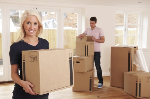 A smiling woman with blonde hair, wearing a dark blue T-shirt, holds a large cardboard box in front of her, standing inside a bright, well-lit room with wooden flooring and large windows that reveal a residential street beyond. In the background, a man in a light pink shirt and dark trousers is seen stacking and organizing additional cardboard boxes of various sizes, some of which are partially open or sealed with packing tape. The room appears to be part of a property in Beckton, featuring neutral walls and a clean, uncluttered space prepared for a house move. Several boxes are placed on the floor and a few are stacked on top of each other, close to the walls, while some items are being carefully carried or organized. The scene captures the typical process of packing and preparing household belongings for a professional removal service, with visible cardboard boxes made of brown corrugated material, some with black printed labels or symbols, reflective of Watson Removals London's expertise in residential moves within East London. The overall environment is calm and orderly, emphasizing the careful handling of personal possessions during a house relocation. The natural daylight enhances the clarity and detail of the scene, highlighting the physical effort involved in the removal process and the residential setting associated with Beckton postcode area.