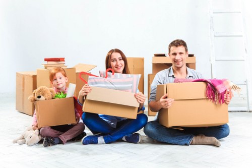 A family of three, including a young girl with red hair, a woman, and a man, sit on the floor indoors in front of a plain white wall. They are surrounded by packed cardboard moving boxes, some open and filled with personal belongings such as a teddy bear, books, and gift-wrapped items. The girl holds a small box containing toys, the woman holds an open box with a wrapped gift inside, and the man is holding a sealed box with a pink scarf draped over the edge. The room has a bright, natural lighting with a clean, minimalist aesthetic, typical of an interior preparing for a house move in Brompton or nearby London. The family appears cheerful as they prepare for their relocation, with the boxes organized in a semi-circle around them. The scene reflects typical household packing activities undertaken by Watson Removals London during a residential move, emphasizing the careful handling of personal belongings in a well-lit indoor space.