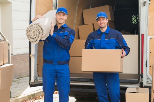 Two professional removal workers dressed in blue uniforms and matching caps are standing outside a residential property, with a removals van parked on the driveway in the background. The worker on the left is holding a rolled-up carpet with a light-colored, textured fabric, while the worker on the right is carrying a plain cardboard box. Behind them, the open rear doors of the van reveal a partially packed interior filled with stacked cardboard boxes of various sizes, some sealed with packing tape, and other household items. The scene is set in a typical suburban street, with the front of a house visible, including a white exterior wall and a brick foundation. The lighting suggests daytime conditions, and the overall atmosphere conveys careful handling and loading of household belongings as part of a household removals or moving service in Neasden. The image emphasizes professionalism and the organized nature of the house removal process, associated with Watson Removals London.