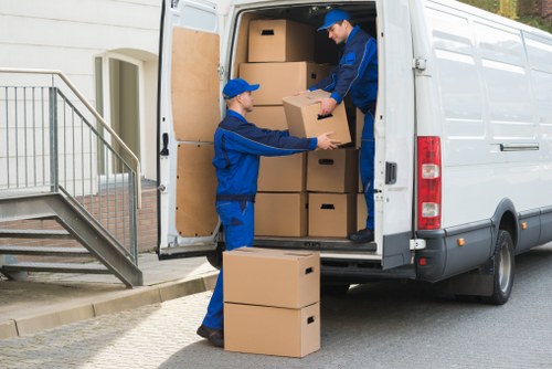 Two movers dressed in blue uniforms are loading cardboard boxes into a white removals van parked outside a residential building with a staircase and a metal railing. One mover is on the ground handing a box to another who is inside the van, stacking additional boxes on top of each other. The boxes vary in size and are plain brown, made of corrugated cardboard, and are stacked neatly within the vehicle. The scene takes place in a suburban area, possibly in London, with natural daylight illuminating the environment. The setting suggests a house move or relocation service, with the focus on careful handling and organisation of household items typical of professional removals by Watson Removals London. The background includes part of a house entrance, highlighting the residential context. This detailed scene emphasizes the logistics involved in house removals in London suburbs or nearby towns, showcasing the company's typical operations.