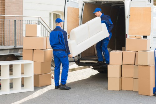 Two professional removalists from Watson Removals London, dressed in blue uniforms with matching caps, are loading a white upholstered sofa into the back of a large grey removals van parked outside a residential property in Barkingside. One removalist is standing inside the van, guiding the sofa, while the other stands outside on the driveway, assisting with the movement. Surrounding the van are several stacked cardboard boxes, some open and some sealed, along with a white plastic shelving unit leaning against the boxes. The setting appears to be on a quiet residential street with a brick house visible in the background, including a small set of stairs leading up to a side entrance. The scene is well-lit by natural daylight, highlighting the organized arrangement of household items prepared for relocation, reflecting the professional house moving services provided in Barkingside by Watson Removals London.