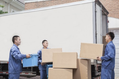 Three male movers dressed in blue uniforms are loading cardboard boxes into the back of a large white removals van parked outside a residential building in Pentonville. One mover is holding a large blue plastic container while another carries a medium-sized box, and the third is handing a similar-sized box to him. The boxes vary in size, with some stacked on the ground and others being moved towards the vehicle. The scene takes place on a paved driveway area, with a brick wall and a grey garage door visible in the background. The lighting indicates daytime, with natural light illuminating the scene, highlighting the neutral tones of the boxes, uniforms, and the van. The environment suggests a professional moving operation, possibly connected to Watson Removals London servicing residents in the Pentonville postcode area during a residential move.