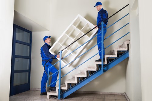 Two professional removals workers dressed in blue uniforms and caps are carrying a white, rectangular bookshelf up a staircase inside a residential building, likely in Mill Hill. The staircase has wooden treads with a natural finish and a blue metal railing, with the risers painted in a darker shade of blue. One worker is positioned at the bottom, gripping the bookshelf with both hands, while the other is at the top, guiding it over the railing. The interior space features white walls, a grey tiled floor, and a glass-paneled door near the bottom of the staircase, suggesting a typical house or flat entrance. The scene appears well-lit by natural or indoor lighting, emphasizing the careful handling of furniture during a move, possibly coordinated by Watson Removals London, operating within the Mill Hill area. The environment indicates careful packing and strategic lifting to protect the furniture and maintain safety during the relocation process.