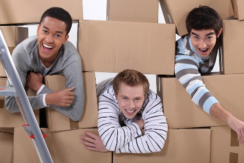 Three young men are inside a room filled with stacked cardboard boxes, which appear to be used for moving or packing. The man on the left, with dark curly hair and wearing a grey sweater, is smiling broadly and leaning on a ladder that is partially visible in the foreground. The man in the middle, with short blond hair and dressed in a white and navy striped shirt, is lying on top of some boxes with his arms crossed in front of him and smiling at the camera. The man on the right, with dark straight hair and wearing a blue and white striped shirt, is leaning forward with one arm extended, appearing to reach or gesture, and has an enthusiastic expression. The boxes behind them are of various sizes, made of brown corrugated cardboard, some with visible tape or packing material. The lighting is bright and natural, suggesting an indoor space, possibly a residential home or apartment in London, as part of a house removal or moving service offered by Watson Removals London, specializing in residential relocations.