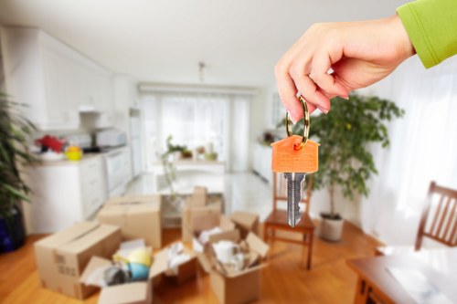 A person's hand, dressed in a green sleeve, is holding a set of house keys attached to an orange keyring, with the keys prominently visible. The background reveals the interior of a residential living space, likely a kitchen and dining area, with many cardboard boxes and packing materials scattered across the wooden floor, some open with contents inside. The kitchen area includes white cabinetry, a countertop with small appliances and utensils, and a window allowing natural light to illuminate the room. There are also chairs, a potted plant near the window, and various household items, suggesting that the scene depicts a home undergoing a move or remodelling. The environment is bright and tidy, with a sense of transition, aligning with professional house removals and relocation services in the Pratts Bottom area within the borough of Bromley, London. The focus on the keys and the cluttered yet orderly scene emphasizes the ongoing process of moving into or out of a home managed by Watson Removals London.