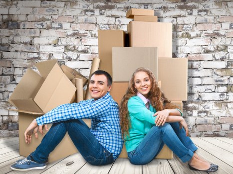 A young man and woman are seated on a light wooden floor in front of a weathered brick wall, surrounded by numerous stacked cardboard boxes of various sizes, most sealed with packing tape. The man, wearing a blue checkered shirt and dark jeans, is sitting with his legs crossed and smiling at the camera. The woman, dressed in a teal sweater and blue jeans, is sitting with her arms folded across her knees, also smiling. The boxes behind them are arranged in a somewhat haphazard manner, with some tilted and open, suggesting an active packing or unpacking process typical of house removal scenes in London or nearby areas. The setting appears to be inside a residential property, with natural lighting that illuminates the scene clearly, emphasizing the textures of the cardboard, the brick wall, and the wooden floor. This image supports a professional removals context at Watson Rremovals London, highlighting household items and moving preparation relevant to local house relocation services in the London postcodes.