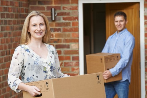 A smiling woman with long blonde hair, wearing a light-colored floral blouse, is standing outside a brick house holding a large cardboard moving box with black printed arrows indicating orientation. Behind her, a man in a light blue shirt and jeans is holding a similar cardboard box, standing near the entrance of the house. The doorway features a wooden doorframe and a partially visible interior leading into the home, with the setting appearing during daylight hours. The background includes a brick wall and a small outdoor wall-mounted light fixture. The scene depicts a house moving or relocation process, consistent with professional removals services in the London area, with the presence of individual items and packing materials typical for household moves.