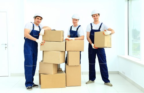 Three professional removals workers dressed in blue uniforms, white shirts, and protective helmets are positioned indoors during a house moving process in Romford. They are standing in a bright, modern room with white walls and large windows allowing natural light to illuminate the space. The workers are holding cardboard boxes of varying sizes, with some stacked on top of each other, indicating an ongoing packing or unpacking activity. All three are wearing white gloves, and two are smiling at the camera while the third has a neutral expression. The foreground features a neat stack of cardboard boxes, some of which are ready to be loaded into a removals van, while in the background the room appears to be empty, with a smooth tiled floor and minimal furnishings. The scene reflects a professional and organized approach commonly associated with house removals services in London, specifically in the Romford area. The setting emphasizes careful handling of household items, including boxes and packing materials, as part of a stress-free moving process provided by Watson Removals London.