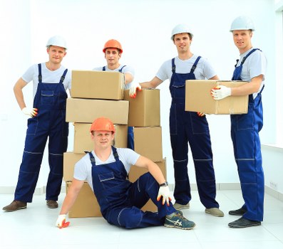 Five professional movers, dressed in blue overalls and wearing protective helmetsâ€”three in light blue, one in orange, and one in a white helmetâ€”pose inside a bright, modern residential property in Whitton, during a house removal process. The movers are handling and arranging cardboard boxes of various sizes, some of which are stacked to form a column, with one mover seated on the floor in front, smiling. The boxes appear to be packed with household belongings, and some are being held or balanced by the movers. The interior environment features a white wall and a large window that allows natural daylight to illuminate the scene, emphasizing the clean and organized setting. The movers are standing or kneeling on a light-colored wooden or laminate floor, with a residential staircase visible in the background. The scene reflects a professional, efficient removal team managed by Watson Removals London, operating in the Whitton area, effectively preparing for a smooth house move.