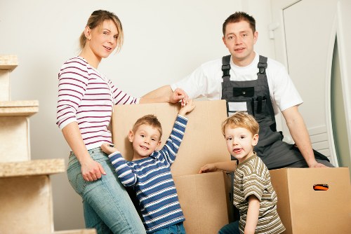 A family of four, consisting of a woman, two young boys, and a man wearing a black work apron, are inside a light-colored house during a move. The woman, dressed in a striped long-sleeve shirt and jeans, is smiling and holding a cardboard box. The two boys, wearing striped tops, are in front of the woman; one is reaching up to hold onto the box while smiling at the camera, and the other looks slightly to the side. The man stands behind them, next to the woman, with one hand resting on the box, and is looking at the camera with a neutral expression. The scene is set near a staircase with beige carpeted steps and a white wall, and the background features a white doorframe. Several packed cardboard boxes of various sizes are stacked around the family, indicating a household move. The lighting is bright and natural, highlighting the details of the furniture and belongings, consistent with home removals in the London area, as serviced by Watson Removals London.