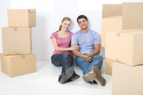 A young couple with light skin, a woman wearing a pink T-shirt and jeans, and a man in a light blue T-shirt and jeans, are seated on the floor of a bright, minimally furnished interior surrounded by packed cardboard moving boxes. The boxes are of various sizes, some stacked and others placed flat on the floor, indicating readiness for a house move in London. The woman has her arm linked with the man's, and they both smile at the camera. The background features plain white walls and an empty space, suggesting a residential property preparing for relocation. The scene is well-lit with natural light, highlighting the texture of the plain cardboard boxes and the casual, relaxed atmosphere of the couple, illustrating a typical moment during a house removals process handled by Watson Removals London, serving the local area and surrounding postcode regions.