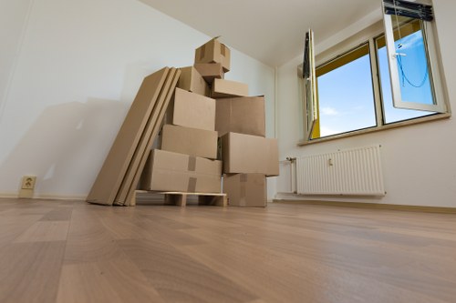 The image depicts a room with light-colored wooden flooring and white walls, featuring a large window on the right side that allows natural light to illuminate the space. In the corner near the window, there is a white radiator mounted on the wall. To the left, there are several stacked cardboard boxes of various sizes, some of which are sealed with packing tape, positioned on a small wooden pallet. Leaning against the wall behind the boxes are several large, flat, rectangular cardboard panels or sheets. The room appears to be in the process of a house move, consistent with professional removals services in the Watson area, as indicated by the clear, unobstructed view of an interior corridor ready for further packing or moving activities typical of Watson Rremovals London. The overall scene emphasizes a clean, uncluttered space prepared for the next steps in the relocation process, with no furniture or personal belongings visible outside the boxes and packing materials, capturing a typical moment of house removal preparation.