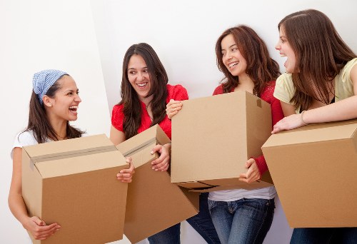 Four women standing indoors in a residential setting, engaged in moving house items, are smiling and exchanging cardboard boxes. The woman on the far left, wearing a light blue headscarf and white top, holds a medium-sized cardboard box, while the second woman, dressed in a red top, accepts a box from her. The third woman, also in red, and the fourth woman, in a yellow top, are each holding larger cardboard boxes, ready for transport. They are standing against a plain, white wall with natural or soft indoor lighting, which highlights their happy expressions. The scene suggests preparation or active involvement in a house removal process, typical of professional removals services in Battersea. Visible items include plain brown cardboard boxes and casual clothing, with no additional furniture or technical equipment shown. The image captures a friendly, collaborative atmosphere characteristic of home moving activities managed by Watson Removals London, providing a relatable visual for those seeking removals services in the Battersea area.