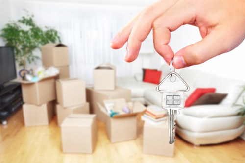 A person's hand holds a silver key attached to a house-shaped keyring, positioned in the foreground of the image. The background shows a cozy living room with a light-colored sofa featuring red cushions, and a collection of packed cardboard moving boxes of various sizes stacked near the sofa. The boxes are plain, brown, and some are open with packing materials visible inside. A green potted plant is situated on the left side, adding a touch of natural decor to the space. The room has wooden flooring and is illuminated by natural daylight coming through large windows, creating a bright and welcoming atmosphere. The scene suggests preparations for house removals in Petersham, with a focus on the visual of key collection and packed belongings typical of professional moving services offered by Watson Removals London, as referenced on their website.