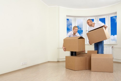 A man and a woman are inside an empty, light-colored residential room with cream walls and a large bay window showing blue sky and clouds. The woman, standing near the left, is holding a medium-sized cardboard box and appears to be preparing to load or unload it. The man, standing on the right, is lifting or stabilizing a similar cardboard box, with several other boxes placed on the light wood laminate floor around them. The scene suggests they are in the process of packing or unpacking during a house removal or relocation in London, as indicated by the context of Watson Rremovals Londonâ€™s services. The room is minimally furnished, with no visible furniture or decor, emphasizing the focus on moving activities. Both individuals are dressed casually, and the space is brightly lit by natural daylight streaming through the large bay window, creating a professional yet practical atmosphere suitable for household moving preparations.