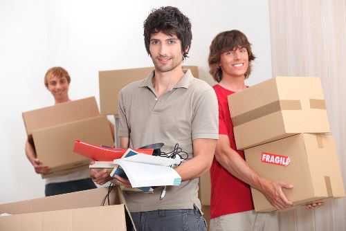 In a brightly lit residential interior, three individuals are preparing for a house move, with a focus on items and packing typical of London-area relocations. The person in the foreground, a young man with dark, curly hair and a light grey shirt, is smiling and holding a red folder, some papers, and a black tape measure, indicating organization and planning for the move. Behind him, on the right, another young man with shoulder-length brown hair, dressed in a red t-shirt, is carrying two large cardboard boxes stacked one on top of the other; the top box has a red and white 'Fragile' sticker, suggesting careful handling. To the left, a third individual with short, blond hair, is holding a medium-sized cardboard box, also part of the packing process. All three are positioned in front of a plain white wall, with parts of the interior visible, such as a window with light-colored curtains. The scene captures typical household items involved in a friendly, organized residential move, aligning with house removals services offered in the Watson area, highlighting the importance of careful packing and use of sturdy cardboard boxes during relocations, such as those by Watson Rremovals London.