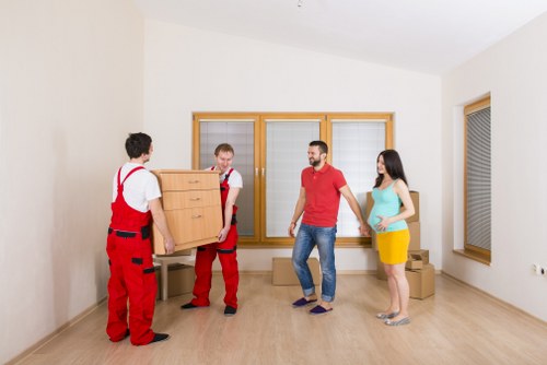 Two removal men dressed in red overalls are carrying a wooden chest of drawers into a bright, minimally furnished room with light-colored wooden flooring and white walls. One man is positioned at the front, supporting the top of the furniture, while the other supports the bottom. A couple, a man in a red T-shirt and jeans, and a woman in a turquoise top and yellow skirt, stand nearby, watching the process. The room features large wooden-framed windows with blinds, bringing natural light into the space, which appears to be part of a residential property in South Kensington. Several cardboard boxes are stacked on the floor in the background, indicating an ongoing move. The environment is clean, spacious, and well-lit, with a residential ambiance suitable for house removals or estate clearance, aligning with Watson Removals Londonâ€™s services in the South Kensington area.