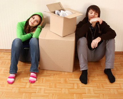 A young woman and a young man are seated on a wooden floor in a room, leaning against a large cardboard box with an open lid containing packing paper. The woman, on the left, is wearing a bright green hoodie, blue jeans, and pink-and-white striped socks, with her head resting on her hand and a neutral expression. The man, on the right, is dressed in a dark brown hoodie, grey trousers, and black socks, with his eyes closed and his hand resting on his knee. The room has plain light-colored walls, and the scene suggests a staging area for a house move, relevant to Watson Removals London's house removals services in London, with the focus on packing and household belongings preparation in a casual, residential setting.