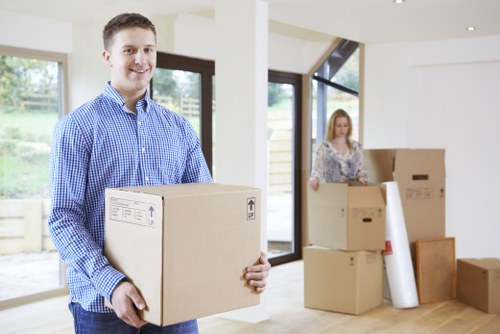 A young man with short dark hair and a cheerful expression is standing in a well-lit, modern room with large windows, holding a cardboard box labeled with shipping or handling symbols. He is wearing a blue checkered shirt and appears to be involved in a house removal or moving process. In the background, a young woman with long blonde hair, dressed in a floral blouse, is standing near a stack of similar cardboard boxes, some sealed and others open to reveal rolled-up packing material. The room has a neutral colour palette with white walls, light wooden flooring, and black-framed windows, offering a view of an outdoor garden area. The scene suggests the process of packing or unpacking during a residential move within the area near Archway, London, as indicated by the context of the website. Watson Removals London is visible as the service provider, supporting stress-free house removals in the local area, with a professional and organized atmosphere centered around household furniture, boxes, and packaging materials.