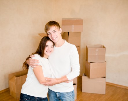 The image shows a young couple with light skin and brown hair, smiling and embracing each other inside a residential property, likely during a house move. The woman is wearing a white short-sleeved top and blue jeans, while the man is dressed in a white long-sleeved shirt and blue jeans. Behind them, there are several cardboard boxes of various sizes stacked and arranged on a hardwood floor, which are ready for packing or unpacking. The background features a plain, beige wall and a section of wooden flooring, suggesting they are inside a living room or entry area of an Aldwych residence. The lighting appears natural or softly artificial, illuminating the scene evenly. This setting reflects typical house removal activity carried out by Watson Removals London as part of their professional removals services, emphasizing the process of packing, preparation, or transition within a home environment in central London. The focus is on the happy, relaxed mood of the couple amidst the moving process, with an emphasis on the household items and the interior space associated with a London-based relocation.
