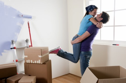 In a well-lit room with white walls and a large window, a man and a woman are engaged in a house removal activity. The man, dressed in a purple T-shirt and jeans, is lifting the woman, who is wearing a blue T-shirt and jeans, off the ground in a joyful embrace. The woman has her arms around the man's neck, and both are smiling. Nearby, there are several cardboard moving boxes, some stacked on top of each other and others placed on the floor, ready for packing or unpacking. A white wall-mounted paper roll dispenser and a red-handled broom or mop are visible on the wall, indicating a clean, organized space with a minimalistic interior. The scene captures a moment of excitement typical during a house move within South Kensington, with Watson Removals London facilitating the transition. The natural daylight coming through the window highlights the warmth and comfort of the interior as the couple shares this cheerful moment surrounded by their belongings in preparation for their move.
