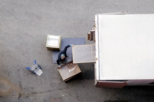 An overhead view of a removal worker inside a white moving van parked on a residential street near Kingsbury, London. The worker is wearing a dark uniform and is loading or unloading a light wood-colored wardrobe with doors slightly ajar, positioned upright near the open side of the van. Surrounding the wardrobe are other household items, including a grey cushioned chair or sofa, a small wooden side table, and several cardboard boxes, one of which is slightly open, revealing contents inside. A blue and white striped shopping bag or storage container is placed on the ground next to the scene, and the ground appears to be asphalt with minor stains or dirt marks. The environment suggests an organized removal process with neatly arranged furniture and personal belongings, indicative of professional house removals services in the Kingsbury area. The lighting is natural, and the scene captures the moment of loading or unloading residential household objects from the vehicle on a typical London street.
