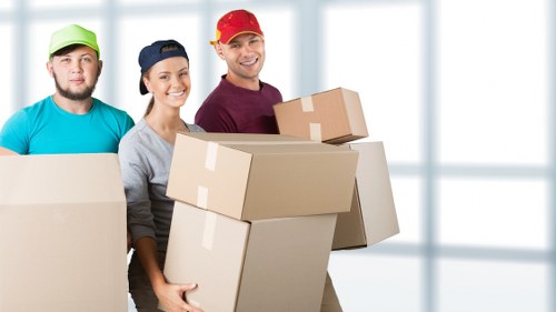 The image features three professional movers from Watson Removals London standing in a well-lit, modern interior space, possibly inside a residential property in Yeading. The movers are dressed in casual work attire with bright-colored caps â€“ one green, one navy, and one red â€“ and are smiling towards the camera. They are holding multiple cardboard boxes of various sizes, which are sealed with packing tape, with visible flaps and corrugated textures. The boxes are in good condition and appear to contain household items for a house move. The background shows a clean, bright room with large floor-to-ceiling windows allowing natural light to fill the space, suggesting an interior ambiance suitable for a professional removals service. The scene emphasizes the process of lifting, carrying, and organizing household belongings, reflecting the expertise of Watson Removals London in residential relocations within the Yeading area. The overall composition highlights the professionalism, readiness, and specific focus on home moving services in the northwest London context.