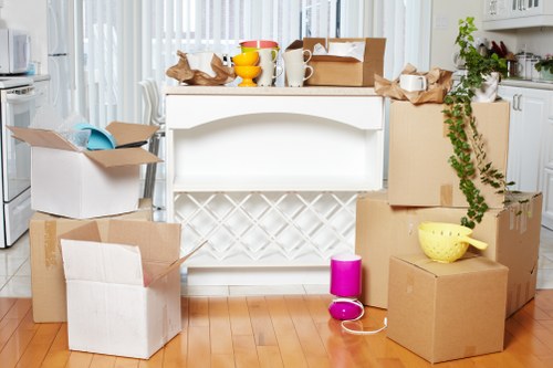 An indoor scene in a residential kitchen, featuring a white kitchen island with a decorative lattice design on its front panel against a background of white cabinetry and a window with sheer curtains. In front of the island, there are several unpacked cardboard boxes, some flattened and others partially assembled, with packing paper and tissue paper spilling out. On top of the island, there are various household items including a brown cardboard box, a yellow bowl, white mugs, and a small yellow container. To the right of the boxes, a pink plastic cup and an electric kettle are placed on the wooden floor, which has a warm, natural finish. A potted green plant with trailing vines is positioned on the countertop to the far right, adding a touch of greenery to the scene. The environment appears well-lit with natural daylight, indicating a bright and airy kitchen space, typical of a domestic setting in Ilford, with a focus on packing and organizing household belongings in preparation for a house move, as managed by Watson Removals London.