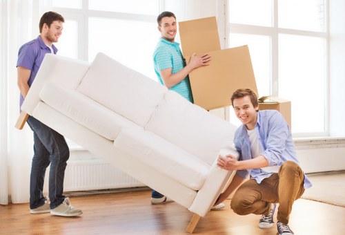 Three young men are engaged in a house removal in a bright, well-lit room with large windows. One man, dressed in a light blue shirt and khaki trousers, is kneeling and steadying a cream-colored fabric sofa with a smooth upholstery surface, positioned in the foreground. Another man, in a purple shirt and dark trousers, is behind him, pushing the sofa from the side. The third man, wearing a turquoise striped shirt and dark trousers, is standing further back, holding a large cardboard moving box and smiling. The scene depicts an active moment of furniture and box transportation within a spacious, tidy room that features wooden flooring, a white radiator beneath the windows, and minimal decoration, consistent with a residential property in Berrylands. Watson Removals London is operating in the background, supporting a professional house move, with a focus on careful handling of household furniture and belongings during the relocation process.