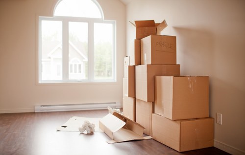 A corner of a room in a residential property shows a stack of brown cardboard moving boxes of various sizes, some taped and sealed, with one open box lying on the light wooden floor, containing packing paper or bubble wrap. The boxes are positioned near a large window with white framing, allowing natural daylight to illuminate the space, suggesting that the room is in the process of being prepared for moving. The wall is painted a neutral off-white color, and a white baseboard runs along the bottom of the wall. The overall scene reflects a typical house removal preparation in Soho, highlighting the early stages of packing within a domestic setting, with no furniture or appliances visible in the image.