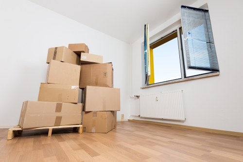 A stack of brown cardboard boxes of various sizes, some sealed with packing tape, are arranged on a wooden pallet in the corner of a bright, empty room with white walls and wooden flooring. The boxes are placed next to a large window with open blue window shutters, revealing a clear sky outside. Below the window is a white radiator mounted on the wall. The room appears to be part of a residential property in London, as suggested by the context of Watson Removals Londonâ€™s website about house moving services. The space is well-lit with natural light, and the room is clear of other furniture or personal items, indicating it is in the process of being prepared for moving or packing. The absence of clutter and the organized arrangement of boxes suggest careful handling typical of professional removals experts. This scene effectively depicts a typical phase during a house removal, where belongings are packed and prepared for transportation within a London home.