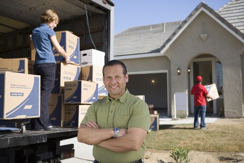 In the image, a professional removalist from Watson Removals London stands in front of a residential house in Covent Garden, with a bright, clear sky overhead. The removalist, wearing a short-sleeved, light green shirt, has a friendly expression and is standing with arms crossed. Behind him, a large moving van with its rear doors open reveals a stack of cardboard boxes, some labeled, ready for a house move. To the left, a young child in a blue shirt and dark pants is seen on the vanâ€™s loading area, handling or arranging some boxes. In the background, a person in a red jacket is walking across the front yard, carrying a smaller box towards the front entrance of the house. The house itself features a light-colored exterior with a tiled roof, a small porch, and external lighting. The scene captures the organized activity typical of a professional house removal in Londonâ€™s Covent Garden area, emphasizing the logistical process of loading furniture, boxes, and household items for a home relocation.