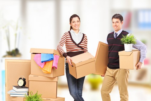 A man and woman are standing indoors in a well-lit room, surrounded by several cardboard packing boxes of various sizes, some open and filled with household items, including a rolled-up pink towel and a blue cloth. The woman, positioned on the left, holds a medium-sized box close to her chest, wearing a striped sweater, jeans, and smiling at the camera. The man, on the right, carries a medium-sized box and is dressed in a dark sweater with a collared shirt underneath, beige trousers, and is also smiling. In the background, there is a blurred view of a residential interior with light-colored walls and large windows, indicating a spacious and modern house environment. On a table to the left, there is a small black alarm clock and some books, while on the right, a potted plant sits atop a cardboard box. The scene suggests a professional house removal service in Aldwych, with Watson Removals London preparing items for a household move.
