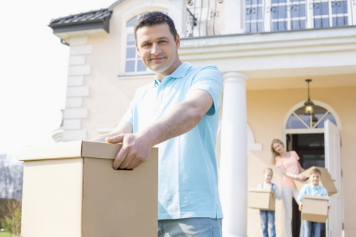 A man in a light blue shirt standing outdoors in front of a residential house with cream-colored walls and white architectural details, holding a cardboard moving box with both hands. Behind him, a woman and a child, both standing on the driveway, are also holding cardboard boxes, suggesting they are involved in a house move. The house features an arched window and a door with a glass panel, with an outdoor light fixture hanging above the entrance. The scene is well-lit with natural daylight, and the background includes a front garden area. This image illustrates residential house removals, typical of services provided by Watson Removals London in the Cambridge Heath area, focusing on professional and stress-free moving activities involving packing and logistics in a homeowners' environment.