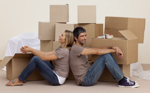 A smiling couple, a woman and a man, sit back-to-back on a beige carpeted floor surrounded by stacked cardboard boxes of various sizes, some open and partially filled. The woman, with shoulder-length blonde hair, wears a light brown t-shirt and dark blue jeans, while the man, with short dark hair, is dressed in a matching light brown t-shirt and blue jeans. They are relaxed and appear to be in a residential living space, with a plain cream-colored wall in the background and a white sheet or protective cover draped over some objects in the corner, indicating a moving or packing process. The scene captures a moment of light-heartedness amid a house removal activity, typical of UK house removals services in London, with no other furniture or household items visible apart from the boxes, emphasizing the packing stage of a house or flat relocation.