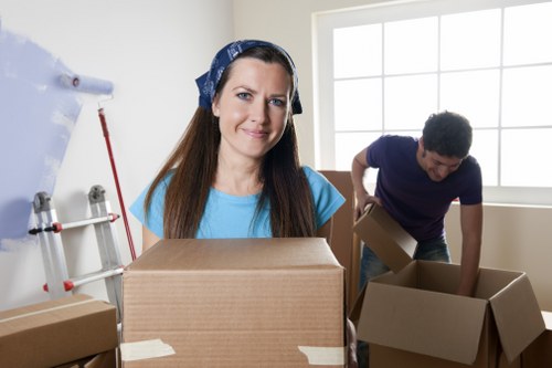 A professional female mover with long brown hair, wearing a blue t-shirt and a patterned headscarf, standing in a well-lit room with large window panes. She is holding a medium-sized cardboard box, sealed with brown packing tape, in front of her. Behind her, a male mover with short dark hair, dressed in a dark blue t-shirt, is seen lifting or arranging another open cardboard box. The room has a white wall, with part of a door or window frame visible, and appears to be a residential interior during a house removal. Several other cardboard boxes, some open and some sealed, are scattered nearby on the floor. To the left, there is a partially visible rack with tools or packaging materials, indicating an organized move. The scene captures a moment of packing and organizing during a house relocation, associated with professional removals services such as those offered by Watson Rremovals London, operating in the London area, possibly within the postal code region associated with the company's service area.