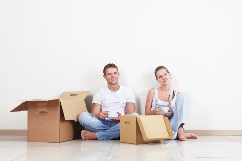 A young man and woman sit on a light-colored tiled floor in front of a plain white wall, surrounded by several cardboard moving boxes of various sizes. The man, wearing a white t-shirt and blue jeans, holds a tablet in his hands and is smiling at the camera. The woman, dressed in a light grey sleeveless top and matching jeans, is also barefoot and sitting with her legs crossed, holding a small stack of documents or papers, and gazing slightly to the side with a neutral expression. Behind them, on the left, there are three open and closed cardboard boxes, some containing household items or packing material. The scene appears to be a residential interior, likely in a house or apartment in London, with a clean, modern environment. This image from Watson Removals Londonâ€™s service page for Payment and Security depicts a typical moment during an upcoming home move, emphasizing organized packing, security, and preparation for a house relocation in the London area.