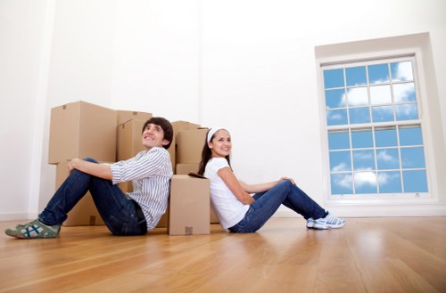 Two young individuals, a man and a woman, are sitting on a polished wooden floor in a bright, empty room with white walls, surrounded by several large cardboard moving boxes. The man, wearing a striped shirt and jeans, is positioned on the left, leaning back against the boxes with a relaxed posture and a smile. The woman, dressed in a white top and jeans, is on the right, also leaning back with her legs extended, smiling towards the camera. Behind them, a large window with multiple panes showcases a clear blue sky with scattered white clouds, allowing natural daylight to fill the space. The scene suggests a house relocation in progress, likely in an area such as Elephant and Castle, with the company Watson Removals London managing the move. The overall atmosphere is cheerful and organized, capturing the moment of settling into a new home before furniture and personal belongings are brought inside.